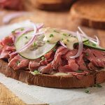 A close-up of a delicious sandwich topped with slices of roast beef, cucumbers, and red onions on dark bread.