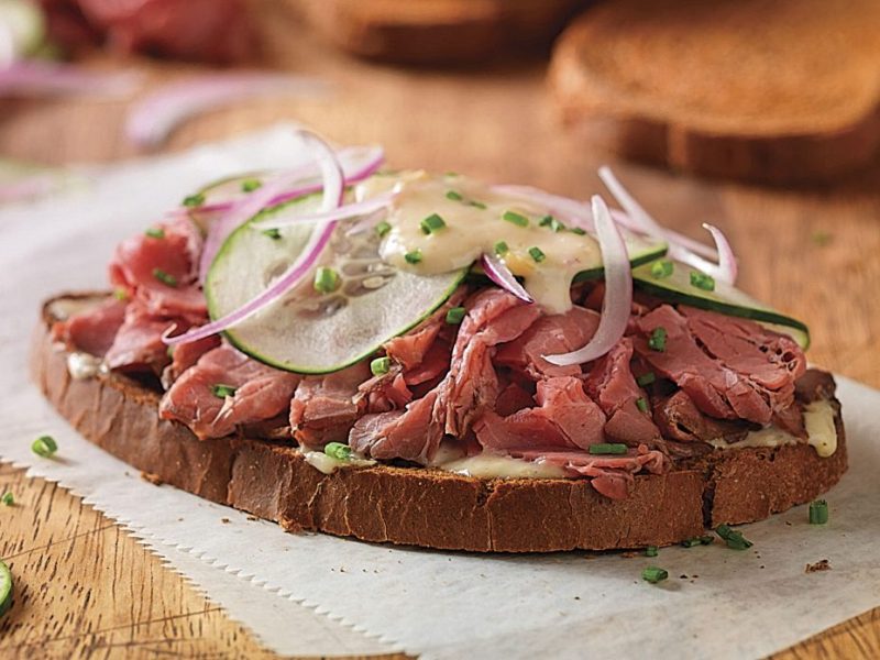 A close-up of a delicious sandwich topped with slices of roast beef, cucumbers, and red onions on dark bread.